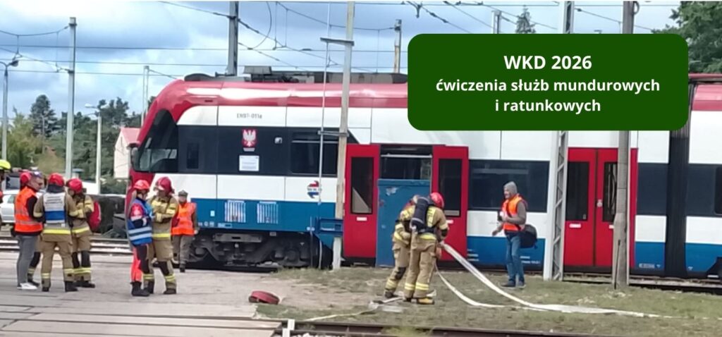 Na zdjęciu widać ćwiczenia służb mundurowych i ratunkowych przy pociągu WKD na terenie stacji lub bocznicy. Kilka osób w strojach ochronnych i kamizelkach odblaskowych pracuje przy wagonie, rozwija węże i prowadzi działania szkoleniowe, a w prawym górnym rogu znajduje się napis „WKD 2026 ćwiczenia służb mundurowych i ratunkowych”.
